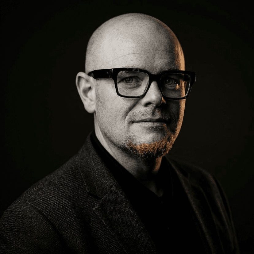 Ryan C. Grulich, wearing dark-framed glasses and a tweed jacket, photographed against a black background.